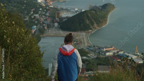 Young man with ponytail descends mountain path and admires view of bay with its port and terminal buildings. Back view.