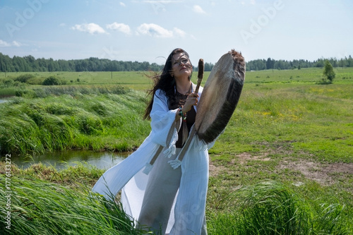 A beautiful female shaman in a trance in the white dress drumming in the natural environment