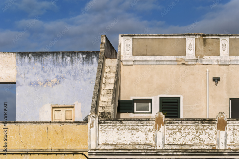 Fototapeta premium Weathered building facade with staircase and small windows, and yellow base with ornate white detailing, all showing signs of age in Olhao, Portugal