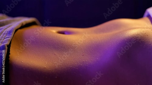 Close up of fit sweaty woman's flat stomach and belly button resting under dramatic vibrant orange and purple light indoors