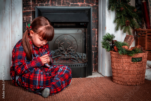 Happy little girl child in pajamas sitting in christmas decorated room near fireplace and prays. Christmas. New Year. St. Nicholas Day. Cozy, festive bedroom interior. Make a wish.
