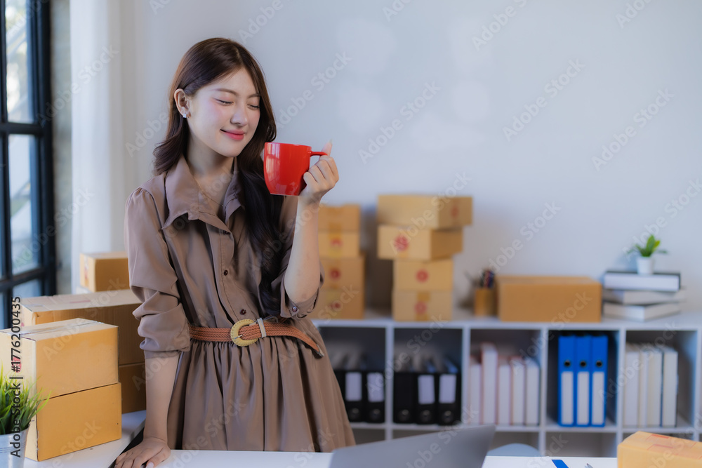 Fototapeta premium Young Asian businesswoman relaxing with a red mug in her home office, managing an e-commerce store surrounded by shipping boxes, laptop, and packaging supplies, smiling contentedly