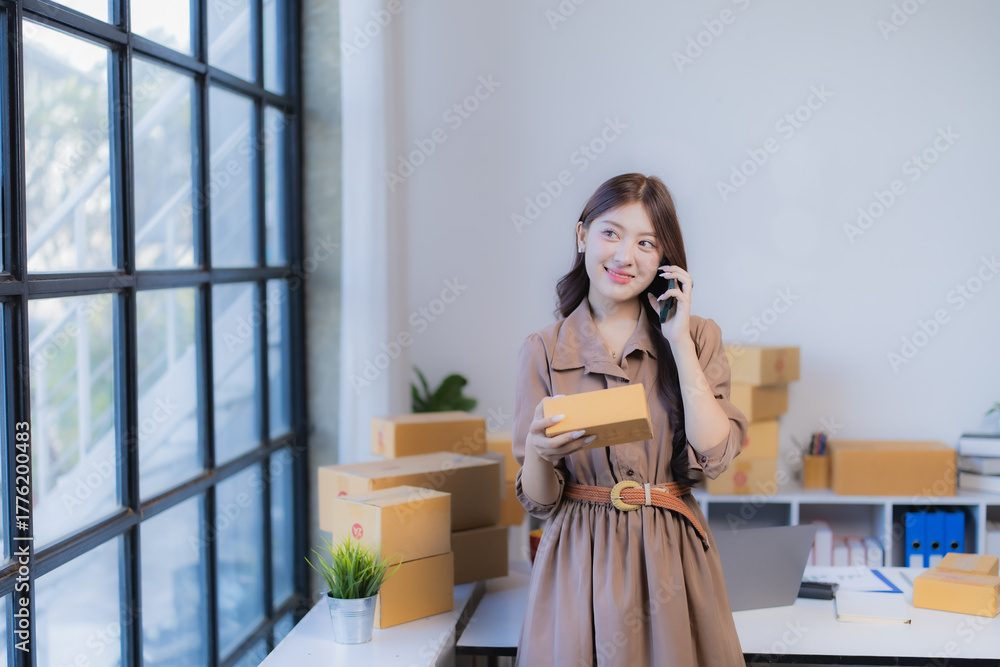 Fototapeta premium Young businesswoman standing in home office, holding a small package and talking on a smartphone, managing online shop orders for e-commerce small business