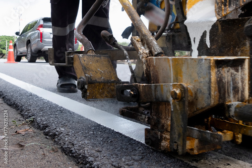 Worker operating road marking machine applying white line on new asphalt pavement. Road maintenance and construction concept, ensuring traffic safety and infrastructure quality.