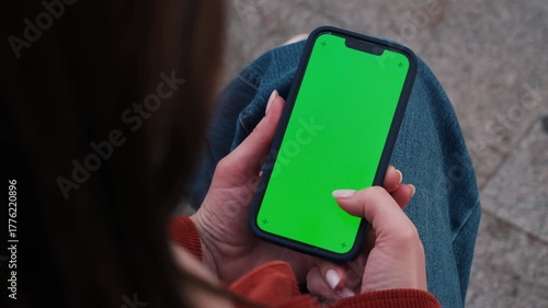 Woman holding smartphone with green screen mockup outdoor, sitting on the bench at the street using phone for chatting with friends, surfing social media, watching reels, video, reading book during re
