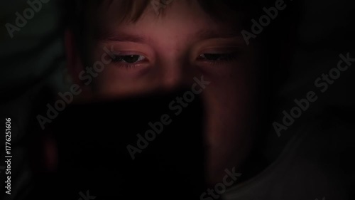 A close-up of a Caucasian teenager's face illuminated by the light of a gadget's screen in the dark, a child using a smartphone in the dark in bed