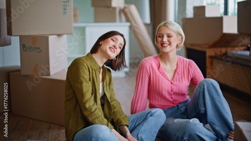 Tired besties moving house sitting living room laughing between carton boxes