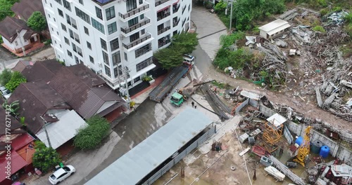 From a high angle, a team of laborers is handling a large pile of recently delivered steel beams within a construction yard that sits immediately adjacent to a modern apartment building