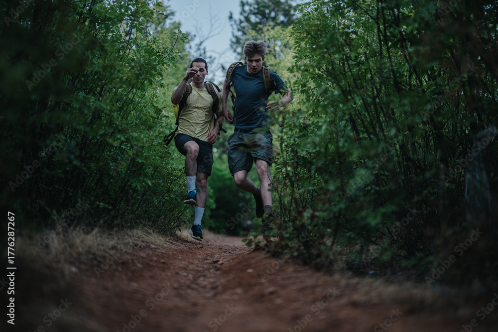 Obraz premium Two men with backpacks sprint along a dirt forest path, capturing energy and teamwork in an outdoor adventure. Ideal for stock listings about fitness, friendship, nature, and exploration.