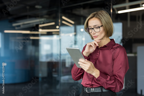 Businesswoman wearing glasses and a maroon shirt standing in a contemporary office, carefully reviewing data on a digital tablet with a focused and contemplative expression
