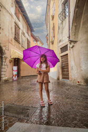 A young girl with an umbrella stands on a rainy day in Modena, Italy, surrounded by wet cobblestone streets, colorful buildings, and the soft reflections of city life in the puddles