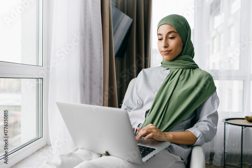 Side view of relaxed young beautiful woman in green hijab or headscarf using laptop in her living-room sitting comfortably next to window and watching series or browsing social media news feed