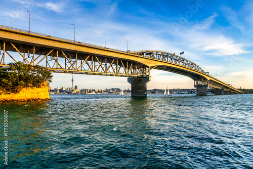 Auckland Harbour Bridge, just as the sun hits the steel structure, with yachts on the water and a view under the bridge to the CBD and Sky Tower.
