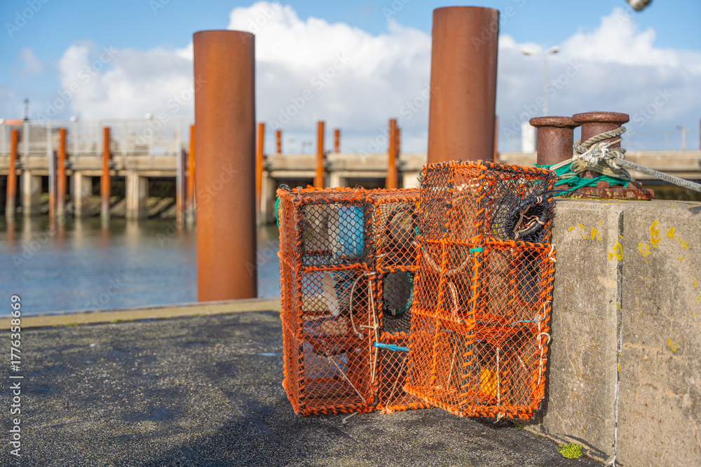 Naklejka premium Two orange fishing cages rest on a pier near rusty poles and mooring ropes. The background shows a harbor bridge and calm sea.