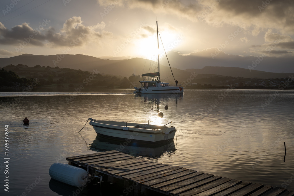 Fototapeta premium A wooden jetty with a small wooden boat, and a holiday sailing yacht in a sea channel with the morning sun rising over the hills in the distance.