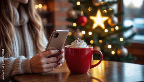 A girl in a coffee shop is holding a smartphone, drinking cocoa with whipped cream.