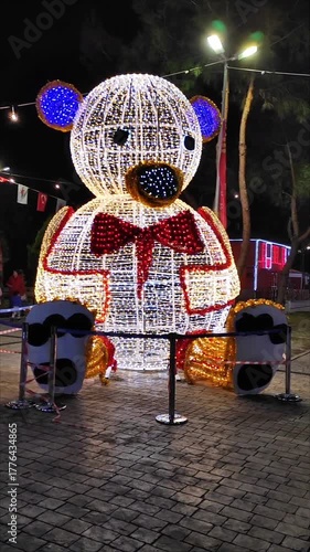 New Year's Eve light installation in the form of a toy bear decorated with lots of bright lights and garlands with blue, gold and red lights on the background of a night city.