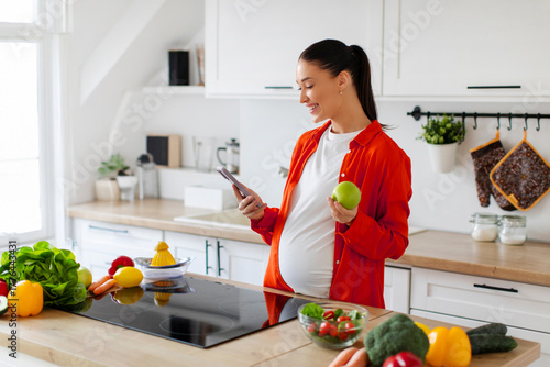 Smiling pregnant woman standing in modern kitchen holding apple in one hand and using cellphone with the other, checking social media feed while cooking or eating