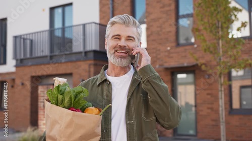 Cheerful Caucasian man with gray hair and beard holding grocery bag full of vegetables talking on smartphone. Standing outside modern residential brick house on sunny day. Remote communication concept