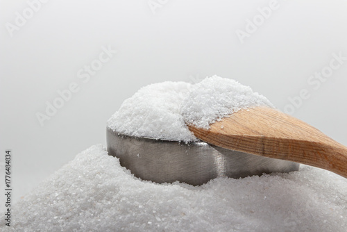 Iodated salt pile is poured into metal container in front of white background, with wooden spoon. Sitting on another pile of salt. Textured horizontal photo, closeup. V14