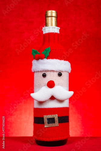 a wine bottle dressed in a festive Santa Claus-themed knitted cover on red at vertical composition