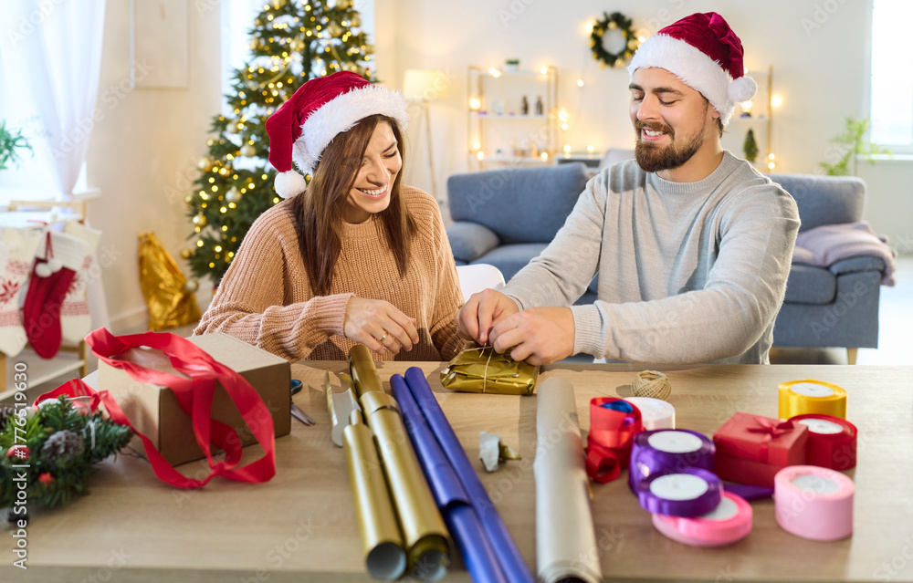 Fototapeta premium Christmas happy couple wrapping gift together near festive tree. In cozy living room, man and woman preparing present box. Love, winter holiday celebration and new year preparation concept.