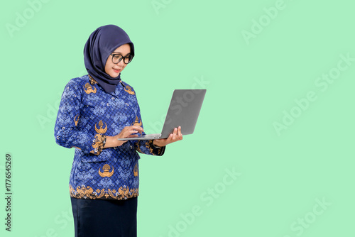 Foto Confident Indonesian Muslim woman wearing blue ASN batik uniform using laptop while smiling, representing professionalism and digital literacy in modern government