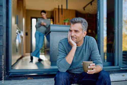 Foto Thoughtful man sitting on porch steps with coffee, smiling, while a woman stands