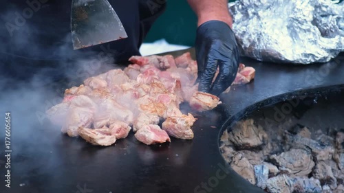 Close-up of a cook using a metal spatula to flip chunks of raw meat on a flat open-fire grill during street food preparation