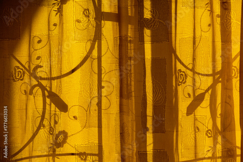 Sunlight casting shadows of an ornate antitheft window grill onto a yellow curtain with floral embroidered pattern