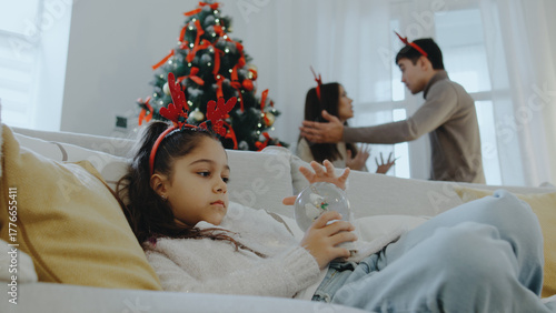 Child with reindeer headband sits on couch while parents argue softly in a cozy living room decorated for Christmas