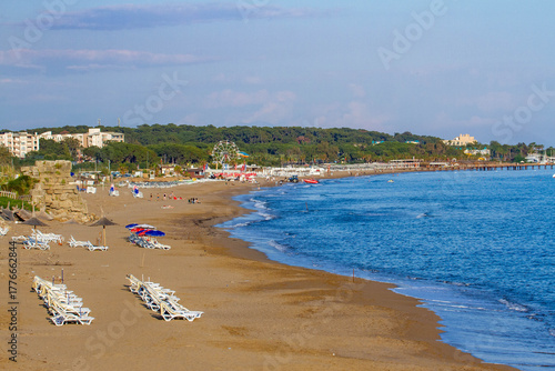 Fototapeta Naklejka Na Ścianę i Meble -  Panoramic view of Side's sandy beach with turquoise water, sunbeds, and sunbathers, backed by the Sorgun forest and resort hotels under a clear evening sky. Antalya, Turkey.

