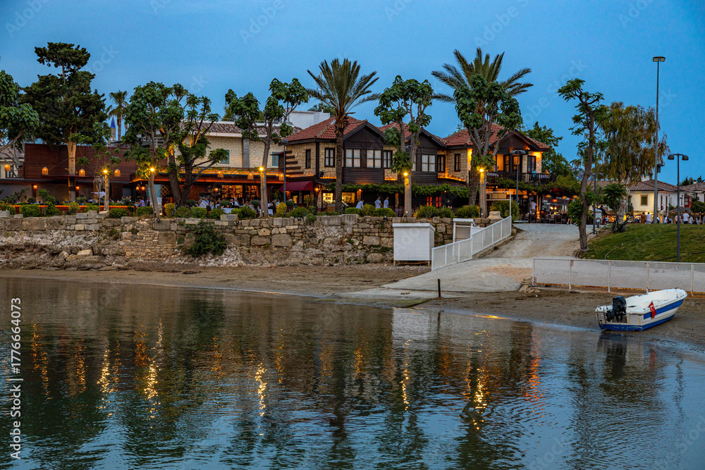 Fototapeta premium A serene post-sunset view of Side's old town from the water, with illuminated restaurants reflecting in the calm sea and traditional stone houses under a deep blue twilight sky. Antalya, Turkey.