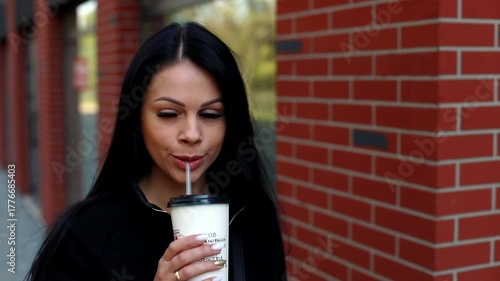 Closeup portrait of smiling attractive brunette young woman drinking through straw looking at camera. Stylish girl walking across the city. Positive lifestyle. Happy day. Healthy nutrition. 