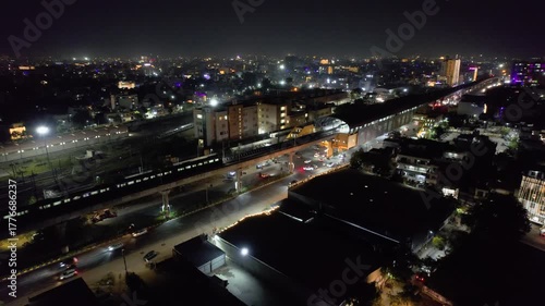 aerial drone descending shot showing metro train arriving at elevated station over busy street and houses in foreground and cityscape filled with lights in the background in indian city of Jaipur