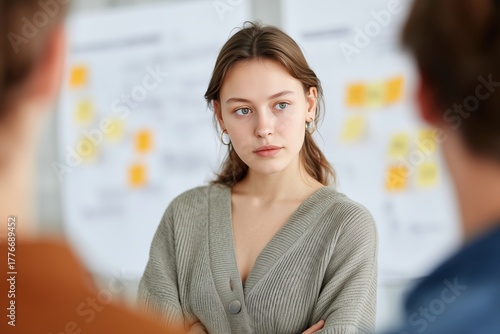 Focused in the Room: A focused young woman stands with folded arms, observing a discussion with colleagues in a professional setting, conveying concentration.