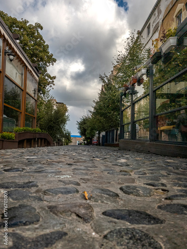 Wallpaper Mural Cobblestone path on Skadarska Street in Belgrade, Serbia, framed by glass cafés and green trees, leading toward a distant light beneath a sky of shifting gray clouds and soft touches of blue. Torontodigital.ca
