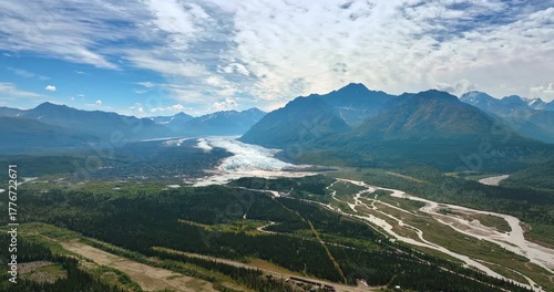 Striking panorama of mountains dazzled by the bright sun. White clouds cover the sky. Branching river flows in the valley. Aerial view. Alaska, USA.