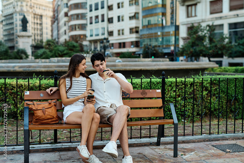 Fototapeta premium Portrait of a happy young couple, woman and man, having fun using a mobile phone and sitting on bench and talking and bonding in park outdoors