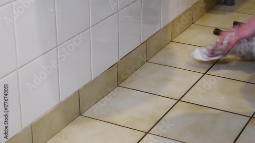 Hardworking person meticulously scrubbing and cleaning the grimy tiles and dirty grout lines along a skirting board with a cloth, performing essential household chores for a clean home