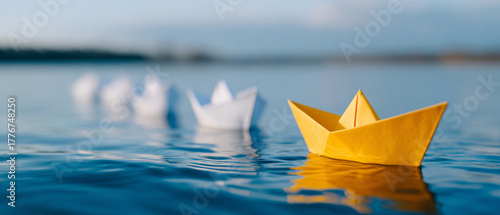 Yellow paper boat leading a group of white paper boats floating on calm water with blurred background and soft natural lighting