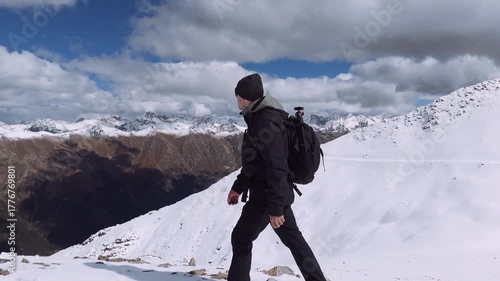 Hiker treks through a snowy mountain area, surrounded by rugged peaks and a vast sky filled with clouds. The scene captures the beauty and isolation of nature on a clear day.