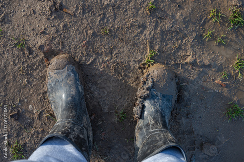 Person wearing black rubber boots standing in wet muddy ground with patches of grass after rain.