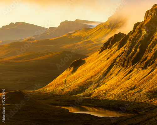 Golden sunrise over Quiraing hills, Isle of Skye, Scotland, captured in warm morning mist. Vertical composition highlighting glowing slopes, depth and peaceful highland scenery.