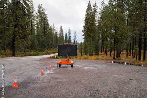 Large, empty digital billboard on a mobile trailer sits in a quiet gravel clearing amid tall pine trees under a cloudy sky, ready for the next message along a remote rural road.