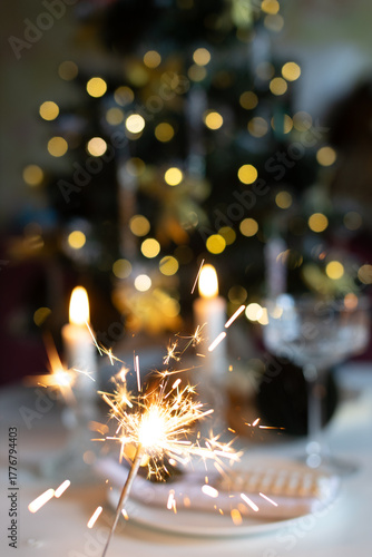 A beautifully set table for a festive event. Two lighted candles, elegant glassware and decorative ornaments are placed near the Christmas tree.