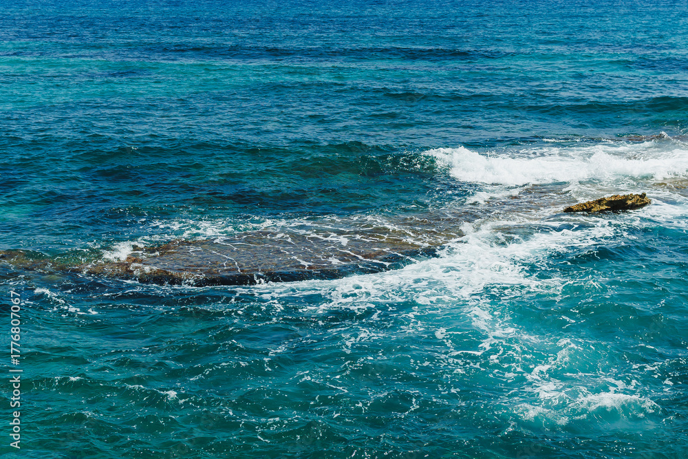 Fototapeta premium Waves crash against rocky shoreline under bright blue sky, capturing the tranquil beauty of the ocean at midday