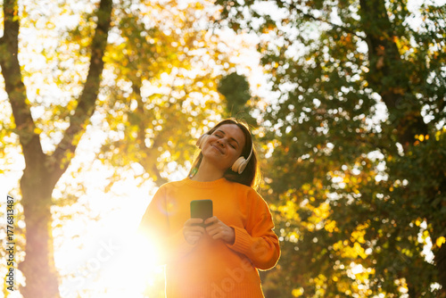 Happy young female using smartphone with wireless headphones in sunny autumn park outdoors