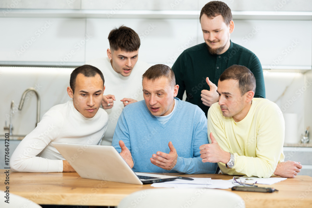 Fototapeta premium Group of men engaged in interested discussion with paperwork and laptop in cozy home kitchen during friendly meeting..