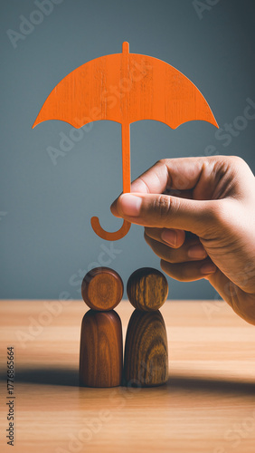Hand holding orange umbrella providing care and security for small wooden figures representing a family, symbolizing protection and insurance
